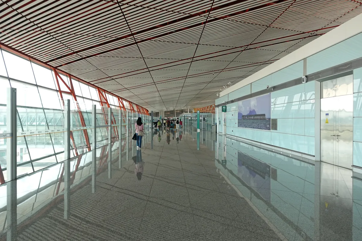 A long, bright arrivals corridor with a red-and-white slatted ceiling and glass walls at Beijing Capital International Airport Terminal 3