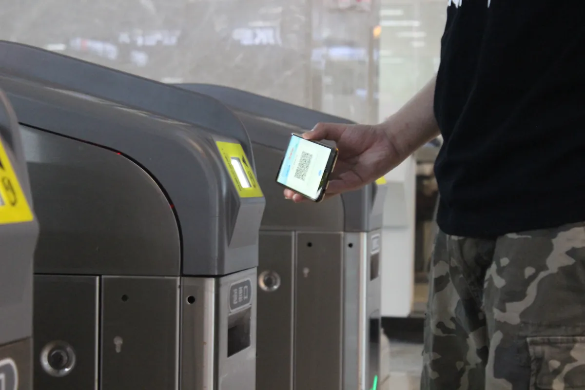 A passenger holds a phone showing a QR ride code against the yellow reader of a metro ticket gate in Wuhan