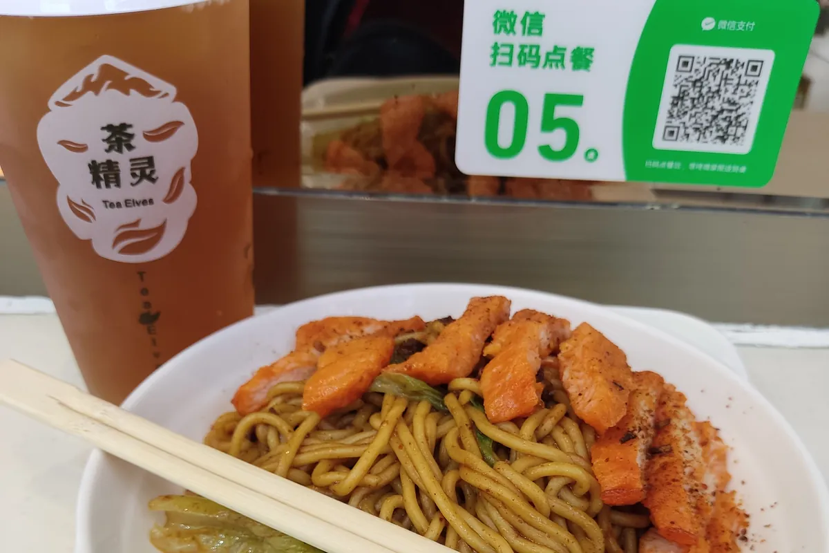 A bowl of noodles with sliced pork chop and greens next to a tea cup, with a green WeChat scan-to-order QR card on the table, at a small shop in Shenzhen