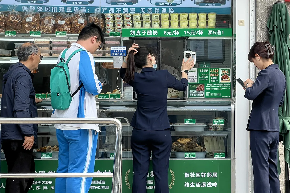 Customers at a street-facing snack shop window in Beijing; a woman holds her phone up to a small payment scanner on the counter while others wait