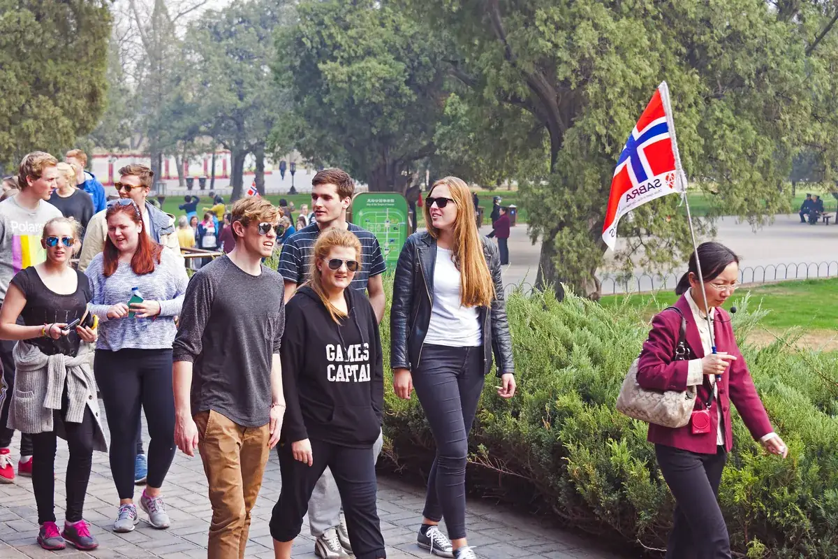 A small group of European visitors walking along a tree-lined path at the Temple of Heaven in Beijing, following a guide who holds up a Norwegian flag