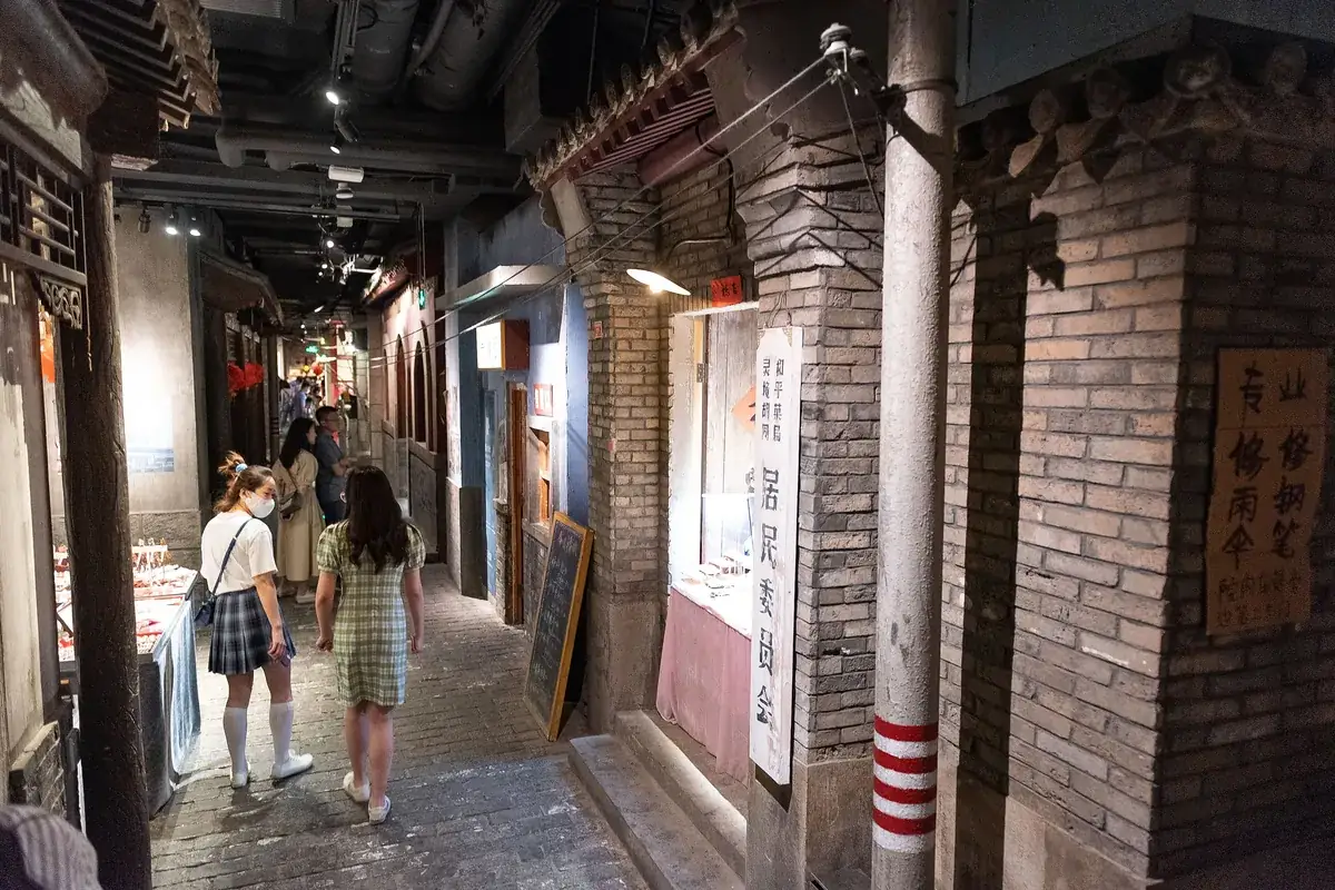 Two young women walking down a narrow recreated old Beijing lane with grey brick walls, hanging lamps and small food stalls on both sides