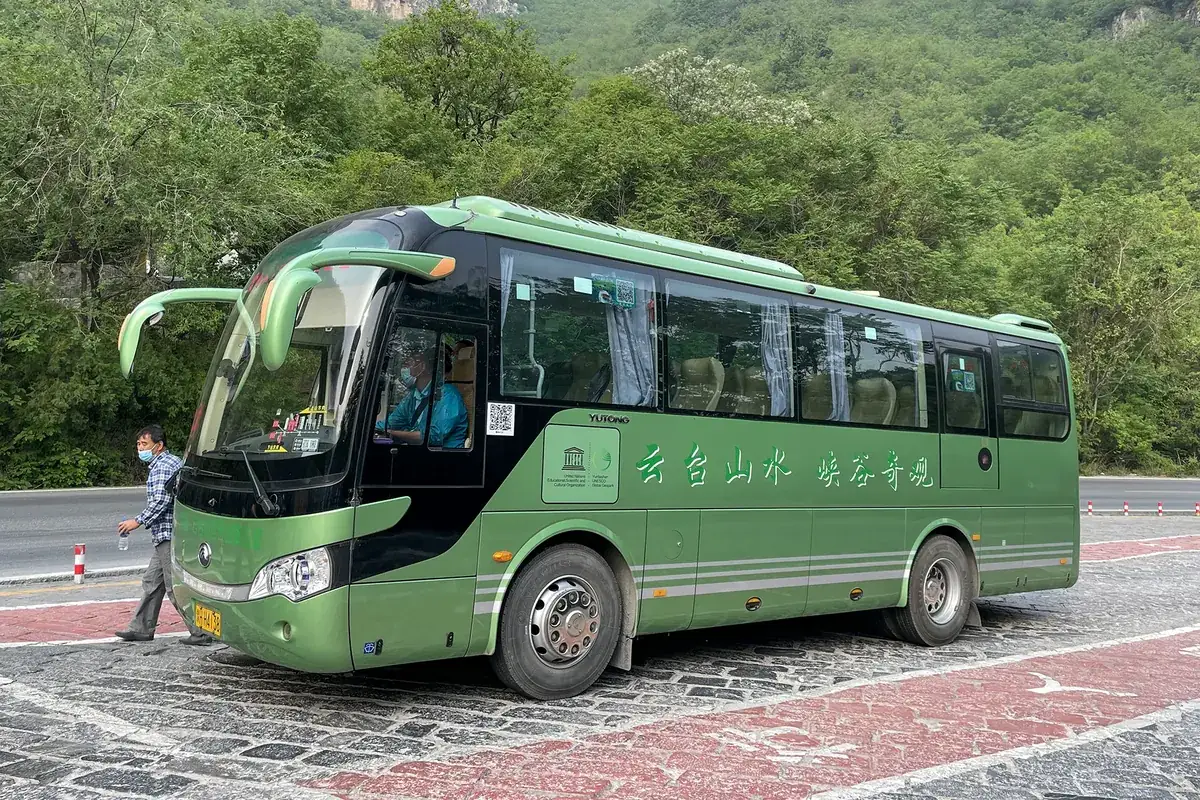 A green tourist coach parked on a paved forecourt in front of a tree-covered hillside, with a man walking past the front of the bus