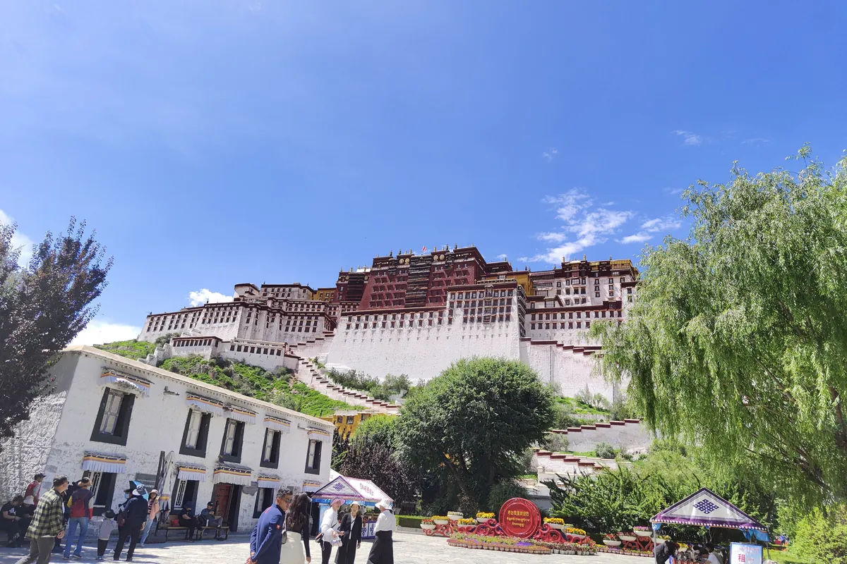 The white and red Potala Palace above trees and a plaza with visitors under a blue sky on a summer afternoon in Lhasa