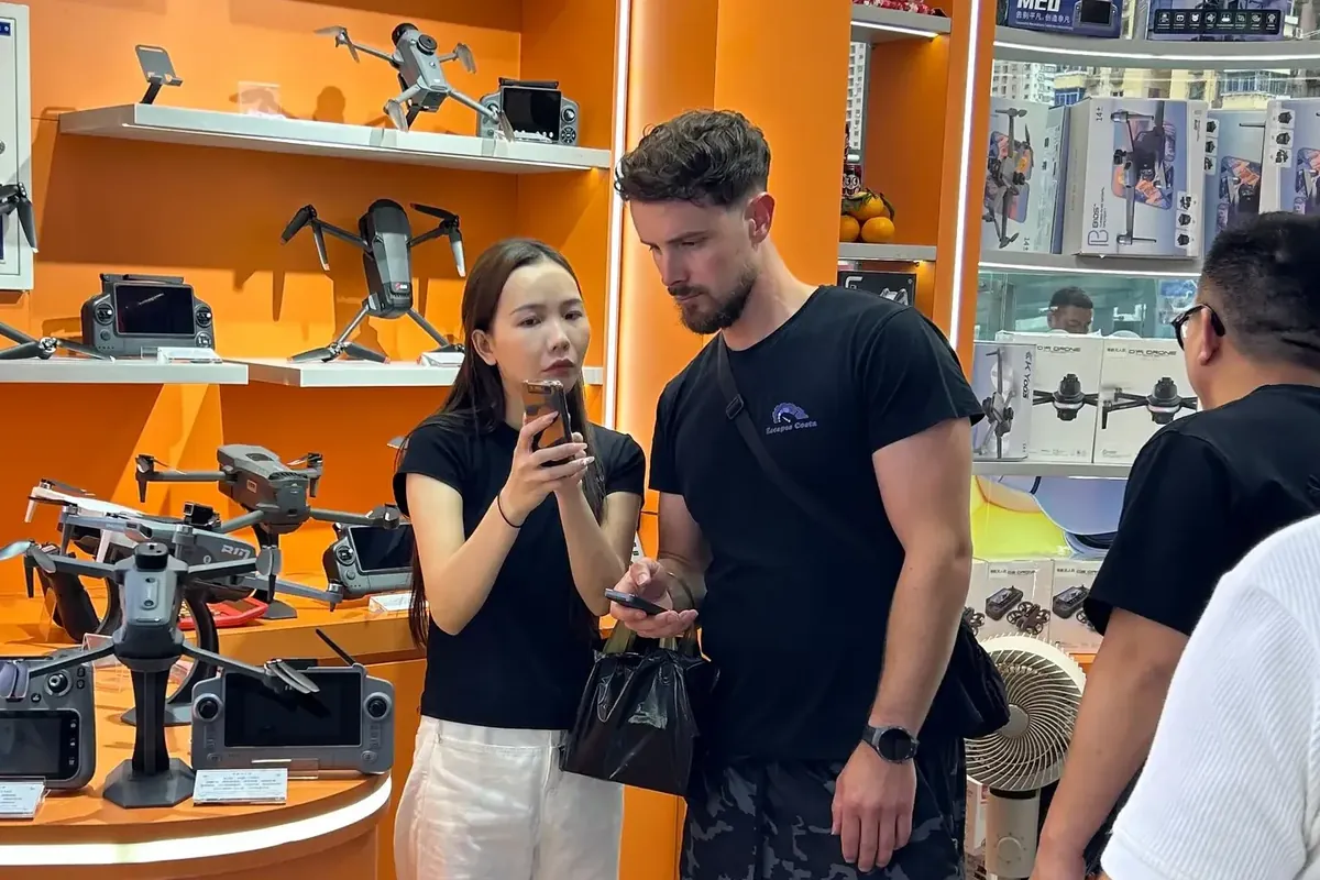 Bilingual escort showing a Western buyer product details on her phone at a drone wholesale counter in Shenzhen