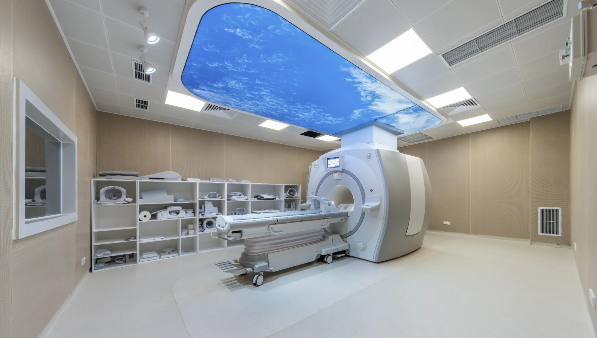 Empty GE SIGNA MRI suite at the Shenzhen partner facility, with a sky-ceiling panel, extended patient table, coil shelves, and control-room window