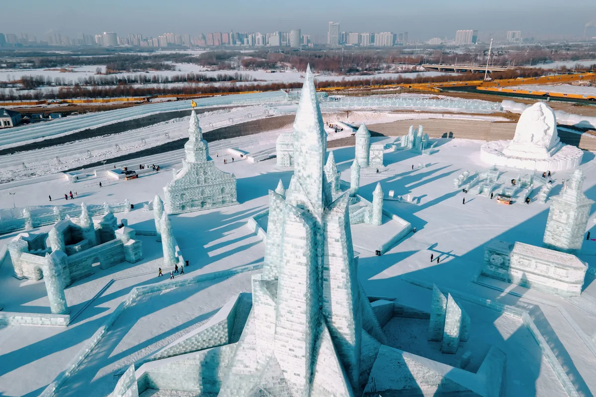 Aerial view of Harbin Ice and Snow World in daylight, white ice towers and castles on the snow with the city skyline behind
