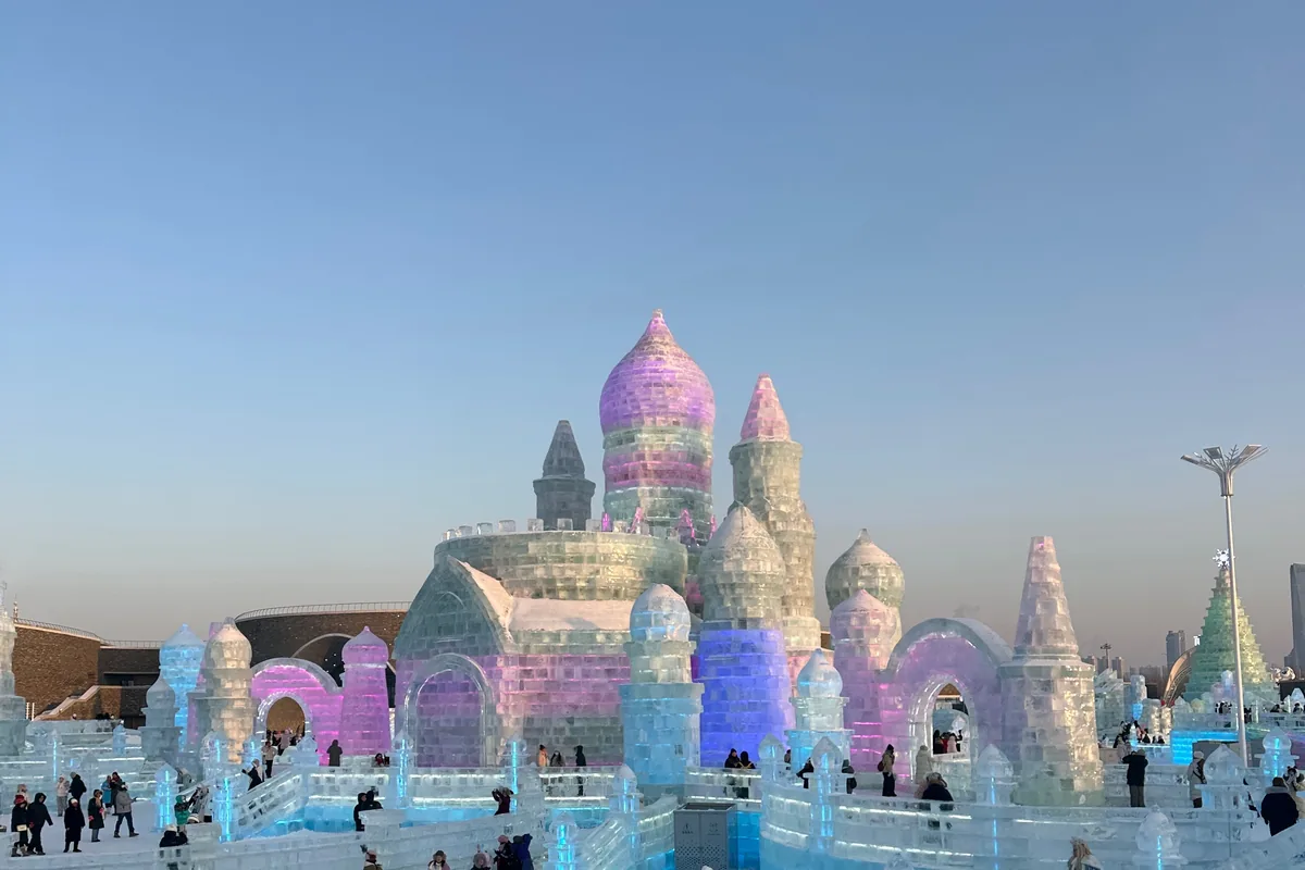 Ice replica of Saint Sophia Cathedral lit pink and purple at Harbin Ice and Snow World in late afternoon, visitors in winter coats in front