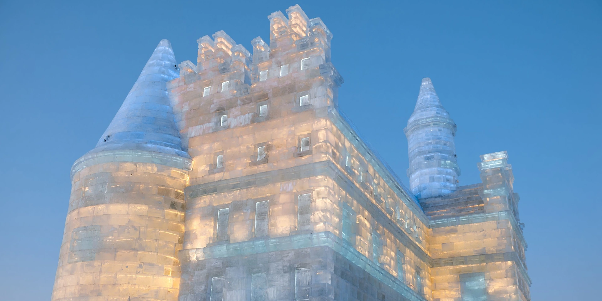Harbin ice castle glowing warm gold from inside at dusk, ice-block towers against a deep blue winter sky