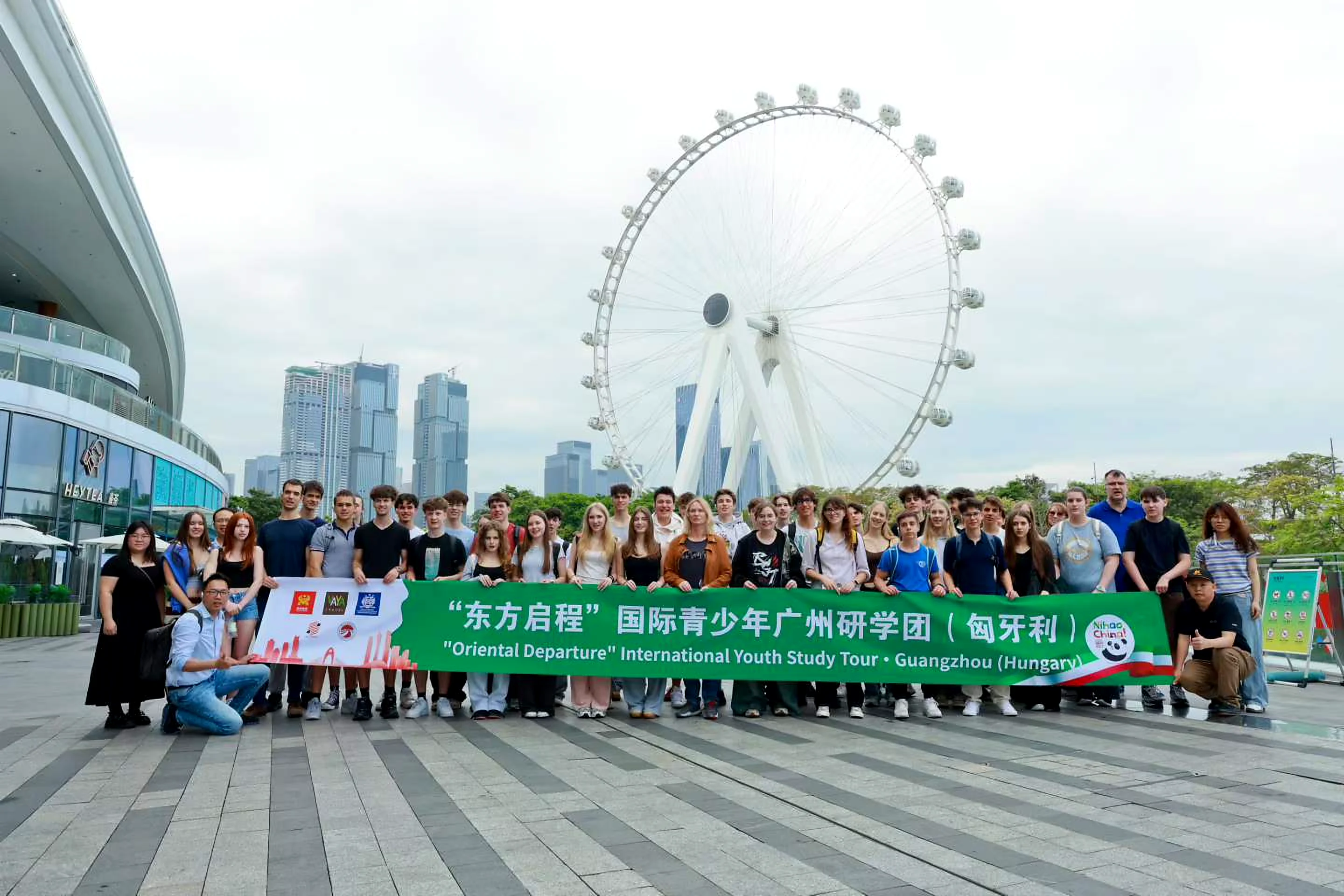 International youth study-tour group with green Oriental Departure Guangzhou (Hungary) banner on a modern Chinese city plaza with a large white Ferris wheel
