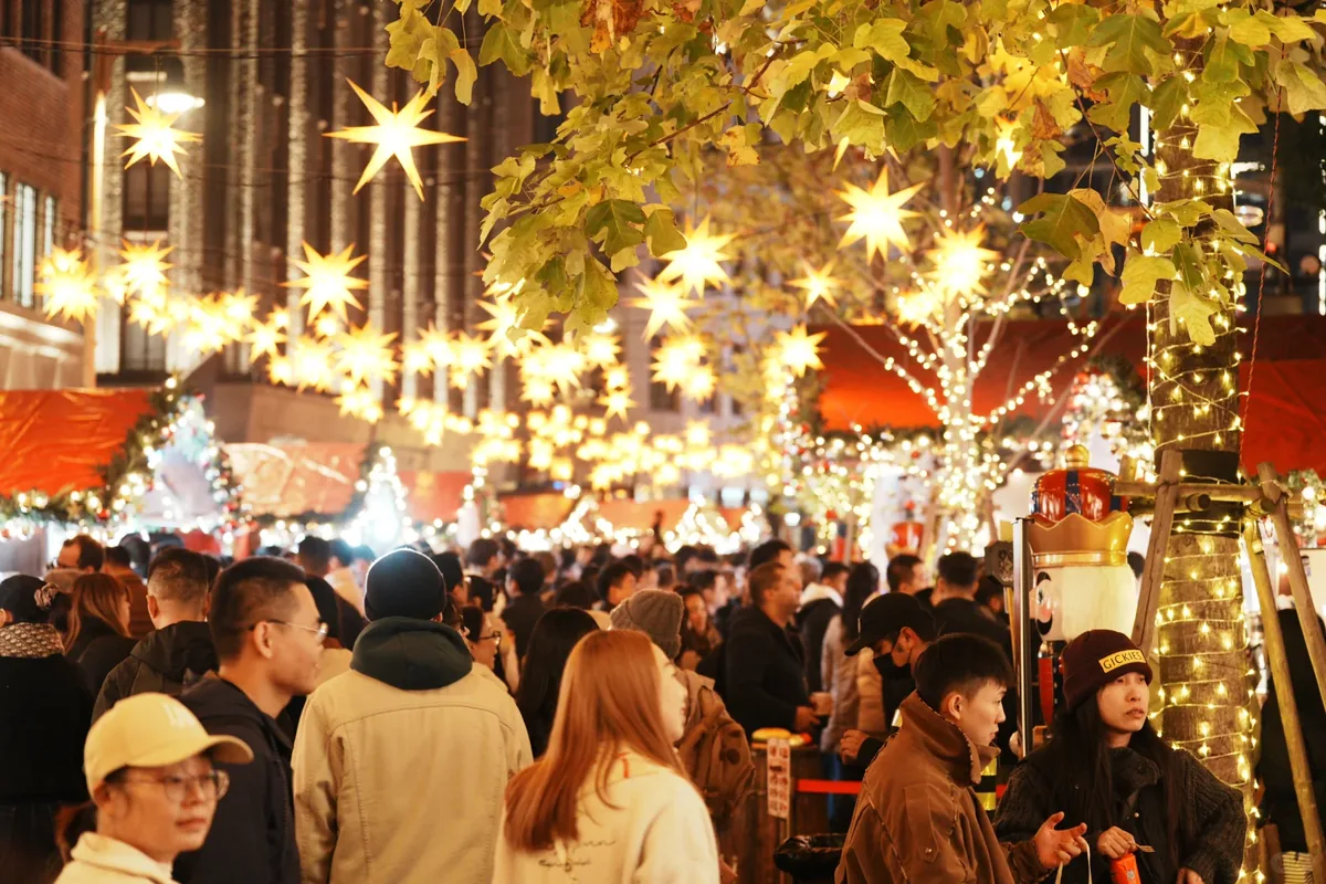 Crowded Shanghai Christmas market walkway under hanging Moravian star lanterns, red-roof stalls, and a large nutcracker figure