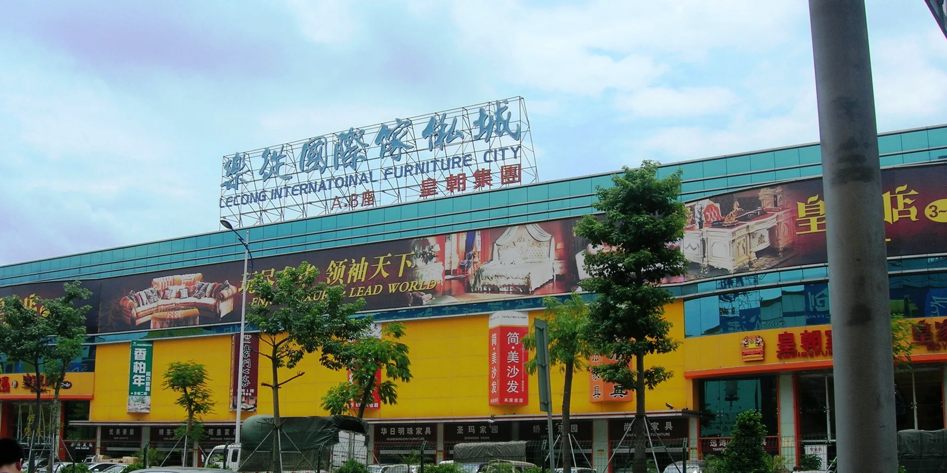 The Lecong International Furniture City building in Foshan, a long yellow and teal mall with a rooftop sign in Chinese and English, furniture billboards along its facade and young trees along the road in front
