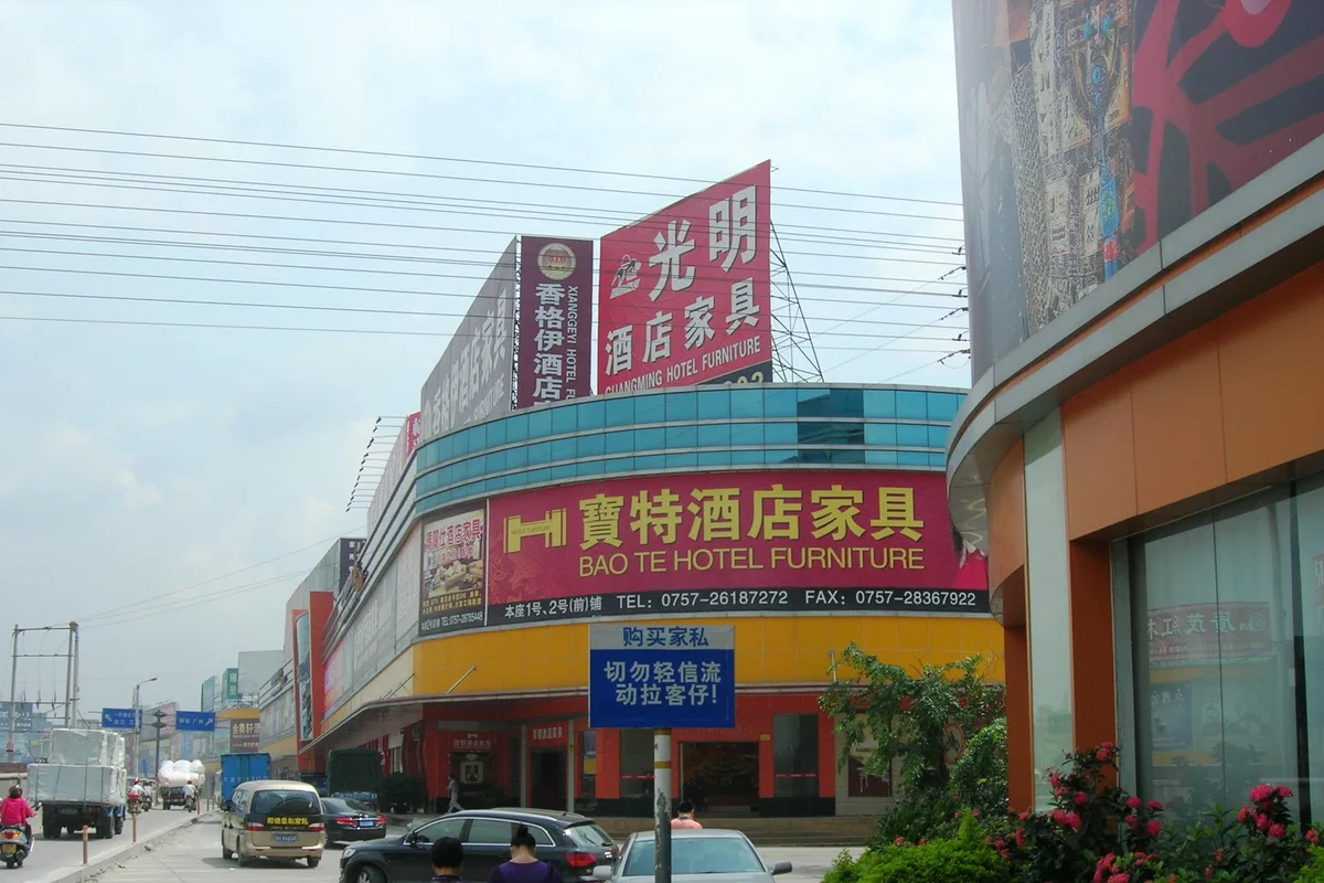A street in Lecong, Foshan, lined with furniture malls, with large red signs for hotel furniture companies above the shopfronts, parked cars and two pedestrians walking in the foreground