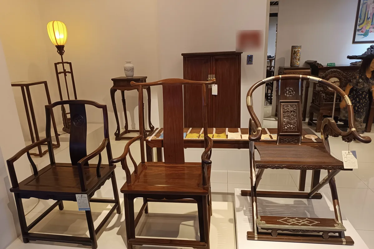 Three Ming-style rosewood armchairs on a white display platform in a Dachong showroom, including a folding horseshoe-back chair with metal fittings, with a cabinet, side tables and a lantern lamp behind