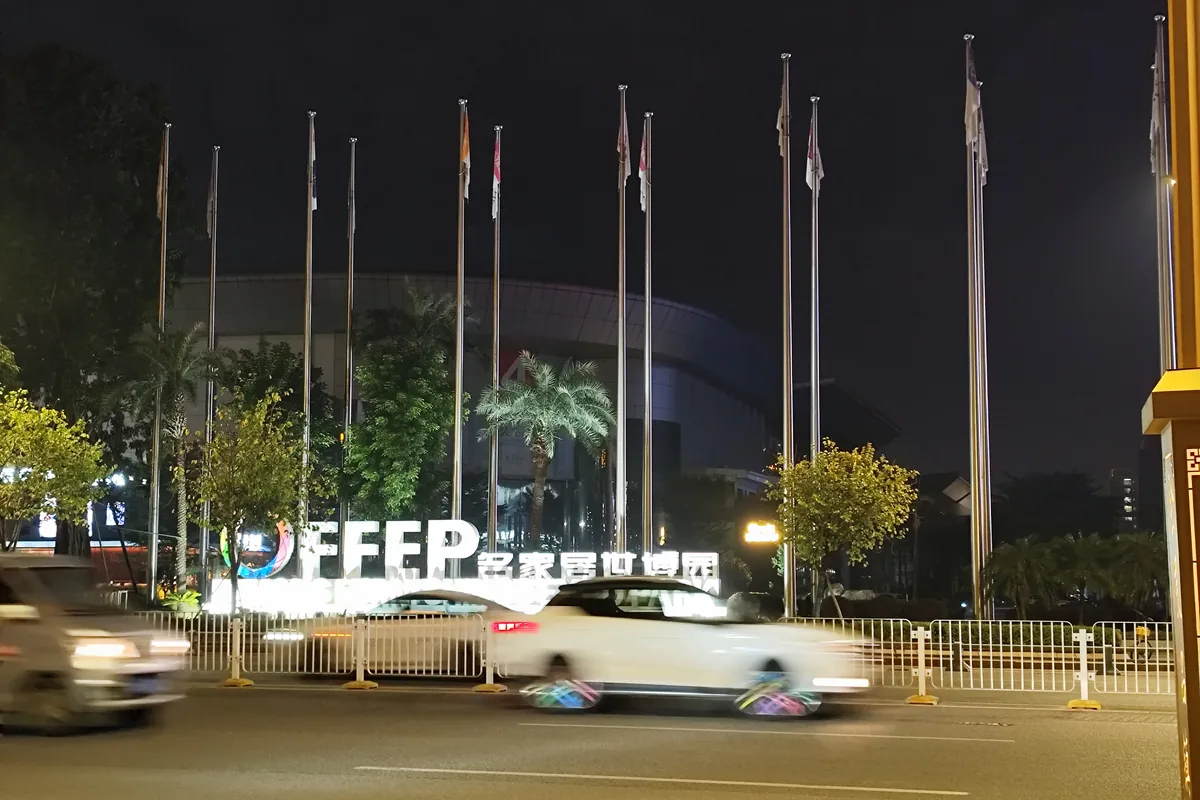 Night view of the illuminated Famous Furniture World Expo Park sign and a row of flagpoles in front of a large curved exhibition building on Furniture Avenue in Houjie, Dongguan, with a car passing in motion blur