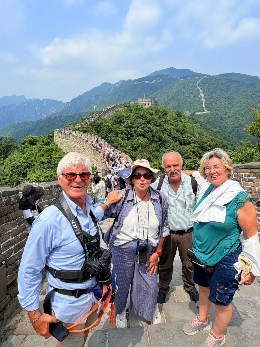 Four travelers posing on a paved Great Wall walkway as the wall climbs tree-covered ridges toward a watchtower