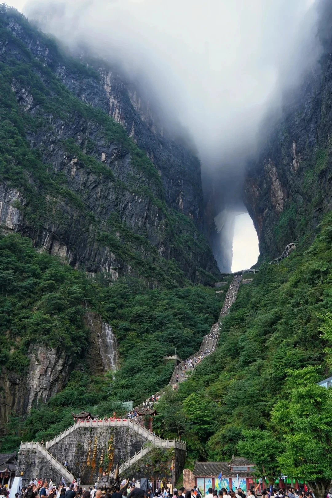 Tianmen Cave (Heaven’s Gate) glowing through mist on a limestone peak, with cliff vegetation and fog at the arch