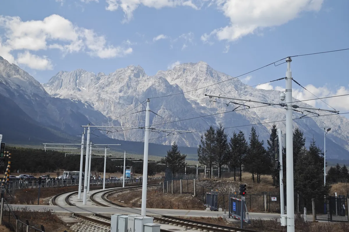 The snow-covered peaks of Jade Dragon Snow Mountain under a blue sky with light cloud, seen across a valley with dark pine trees and overhead tram lines in the foreground