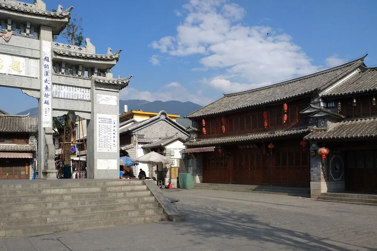A quiet stone-paved square in Xizhou with a carved memorial archway on the left and two-storey Bai wooden houses under a clear blue sky