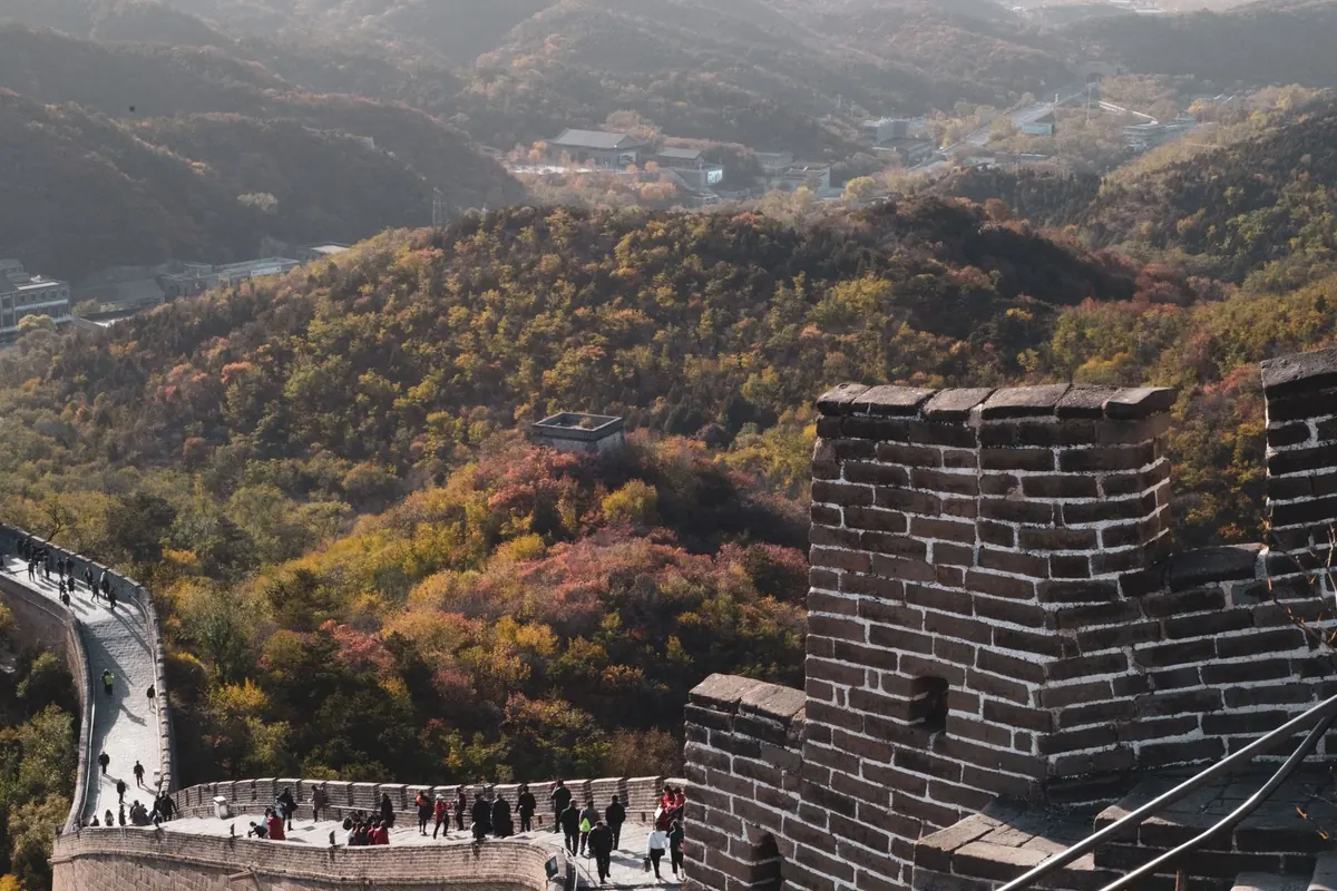 Visitors spaced along the Great Wall of China in autumn, the wall curving through wooded hills above a watchtower