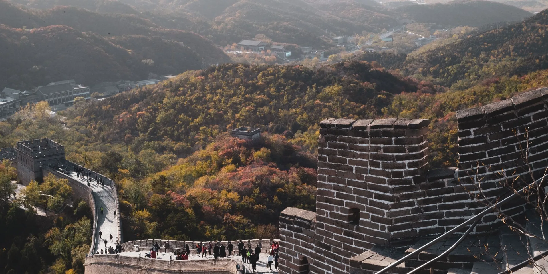 Visitors spaced along the Great Wall of China in autumn, the wall curving through wooded hills above a watchtower