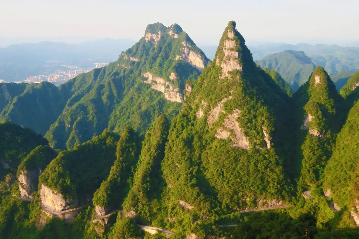 Switchback mountain road below green limestone peaks at Tianmen Mountain, Zhangjiajie, with a town in the far valley